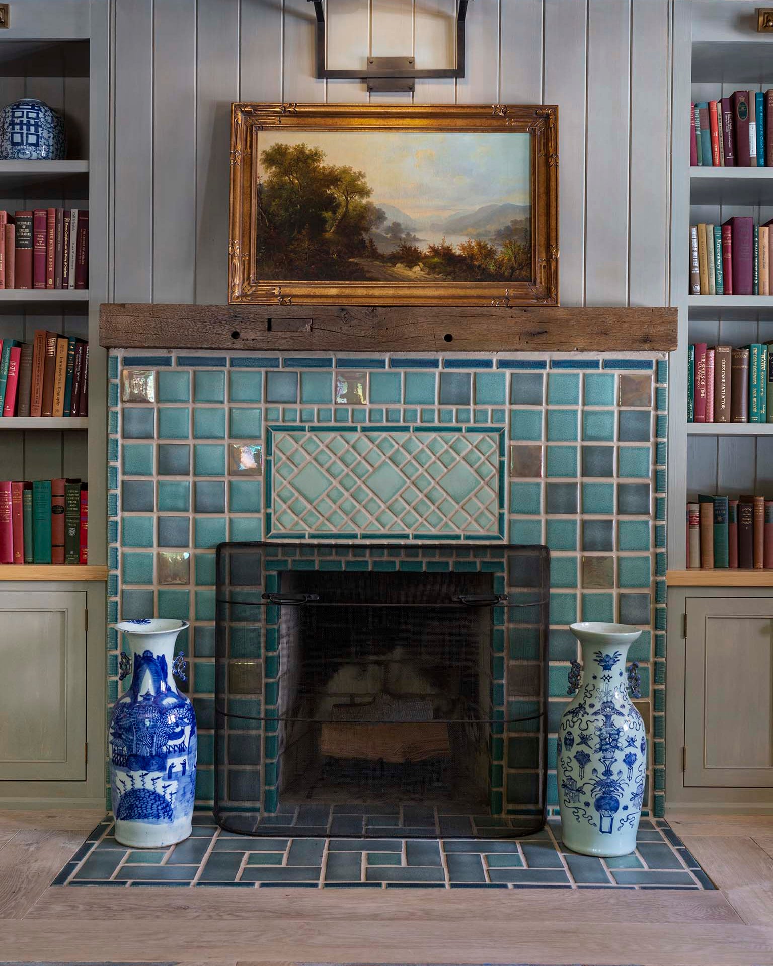 A fireplace with blue, green, and beige tiles, featuring architectural glazes, with bookshelves in the background and decorative blue and white vases on either side of the fireplace.