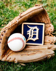 A leather baseball mitt is nestled in bright, green grass. Inside the mitt lays a baseball and our 4x4" Tigers Old English "D" Tile in a Navy Blue and light   "Natural" clay border. The Old English "D" logo is also outfitted in the light clay body, allowing it to stand out against its bright backdrop.
