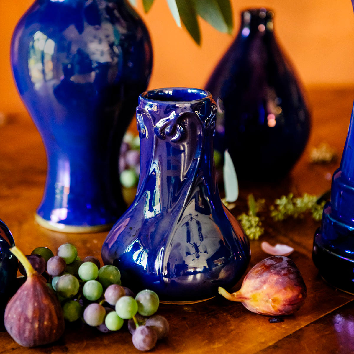 A handcrafted Azurite iridescent Pewabic Snowdrop vase is displayed on a table alongside fruits and in a setting that includes other Azurite vases in the background.