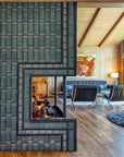 Side view of the lit fireplace. Two mid-century modern chairs and a cozy living room area are visible in the frame. The natural-finished wood vaulted ceiling is a beautiful contrast to the strong geometric design of the fireplace installation.