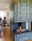 Two women converse at a kitchen island over a cup of tea. The custom three-sided fireplace is in view. From this perspective, the Iridescent tile details and featured Scarab motif are very visible. The fire is lit, creating a cozy and inviting atmosphere. 