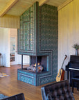A quarter view of a three-sided custom Pewabic Tile fireplace with a strong geometric pattern of subway and custom-cut tiles. The front of the fireplace prominently incorporates our signature Iridescent Aurora glaze with a Matte Green Iridescent Scarab Paperweight at the center. There is a wall piano in the corner and the fireplace is set in a spacious mid-century modern ranch home. 