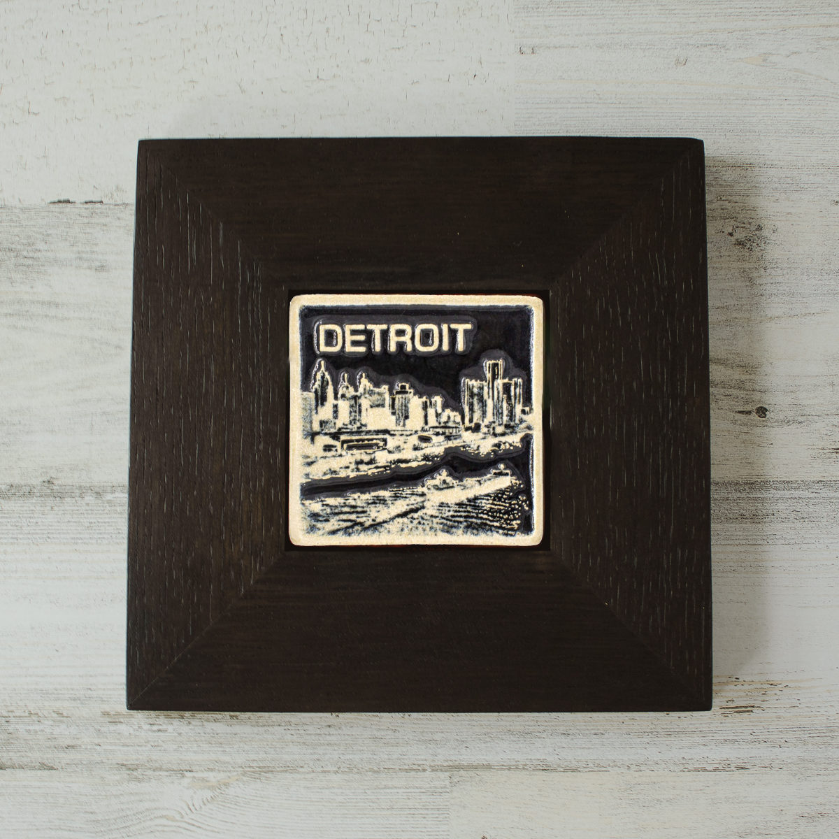 The Detroit Skyline tile features the current downtown skyline with the Detroit River in the foreground. A freighter floats by causing ripples on the otherwise flat surface of the water. The word "Detroit" is written above in the sky. The Skyline buildings, freighter and word are glazed in a glossy white while the background is glazed in a deep black. The wooden frame is matte black.
