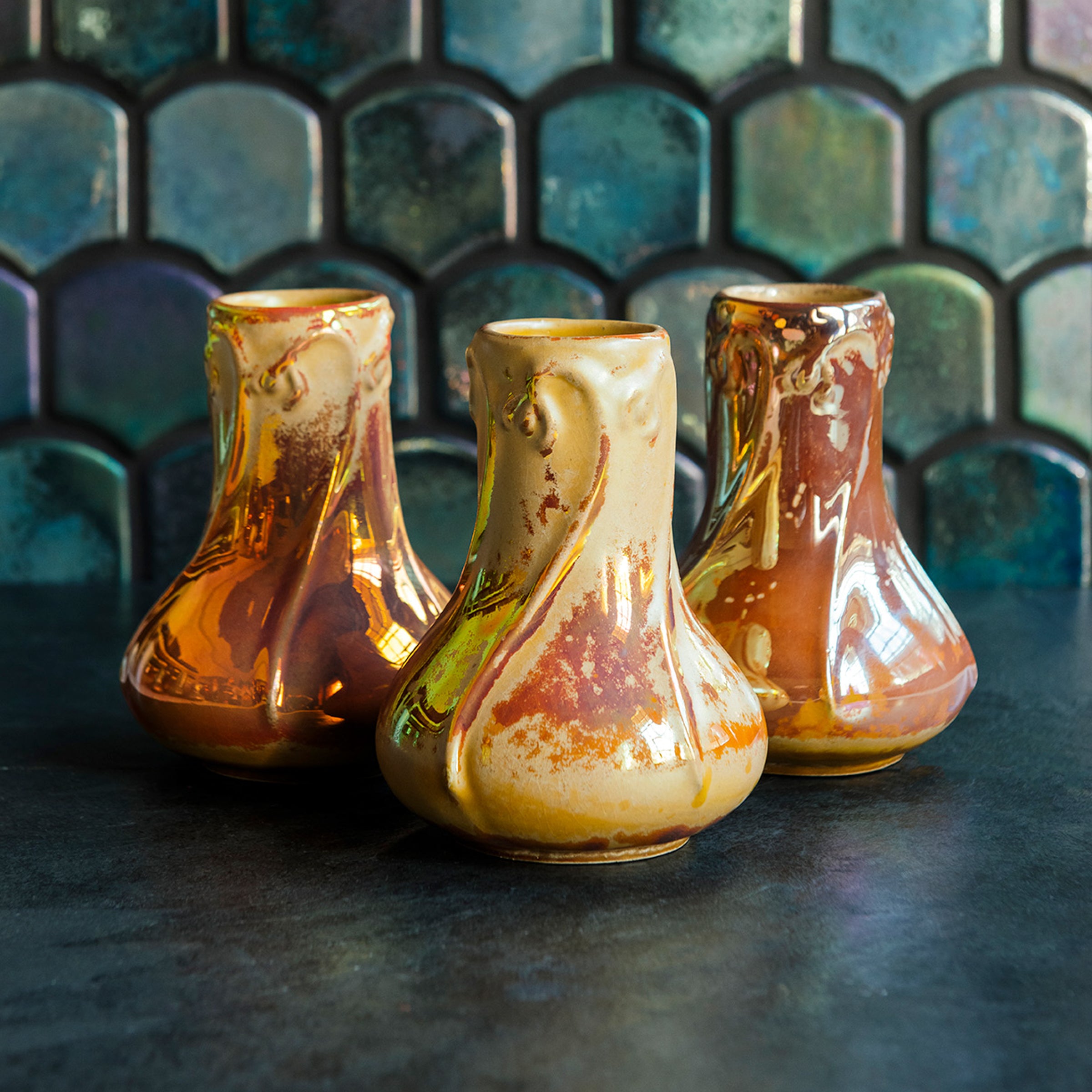 Three Snowdrop vases in Sunstone iridescent, with a marbled design on a dark slate surface, set against a patterned green tile background.