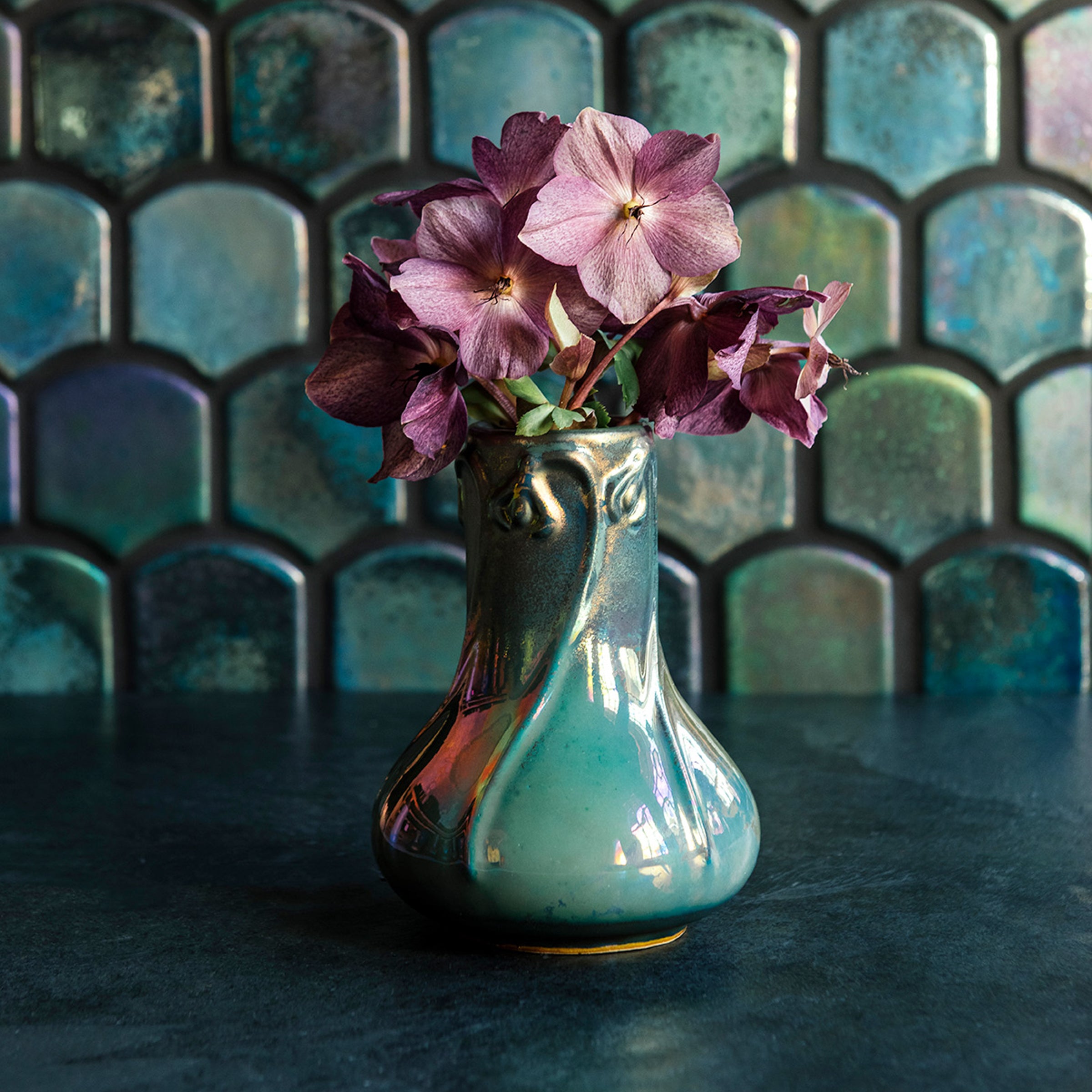 A Matte Green Iridescent Snowdrop vase with hellebore flowers on a dark slate foreground and green, tilled background.