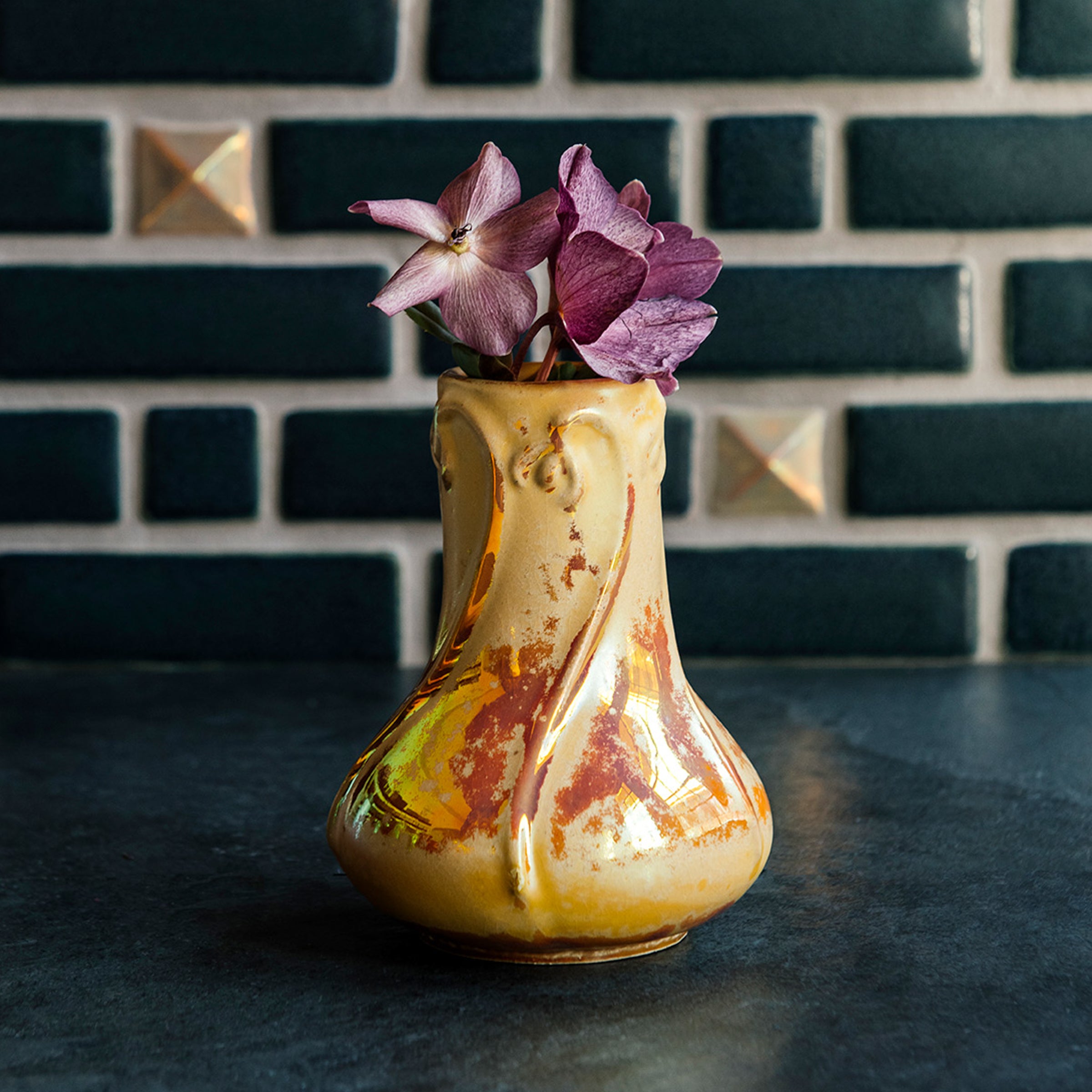 Sunstone Snowdrop vase with purple hellebore flowers on a dark slate surface against a Pewabic tiled wall.