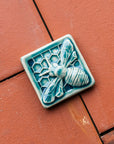 A glossy blue bee tile rests on deep terracotta colored roof tiles. 