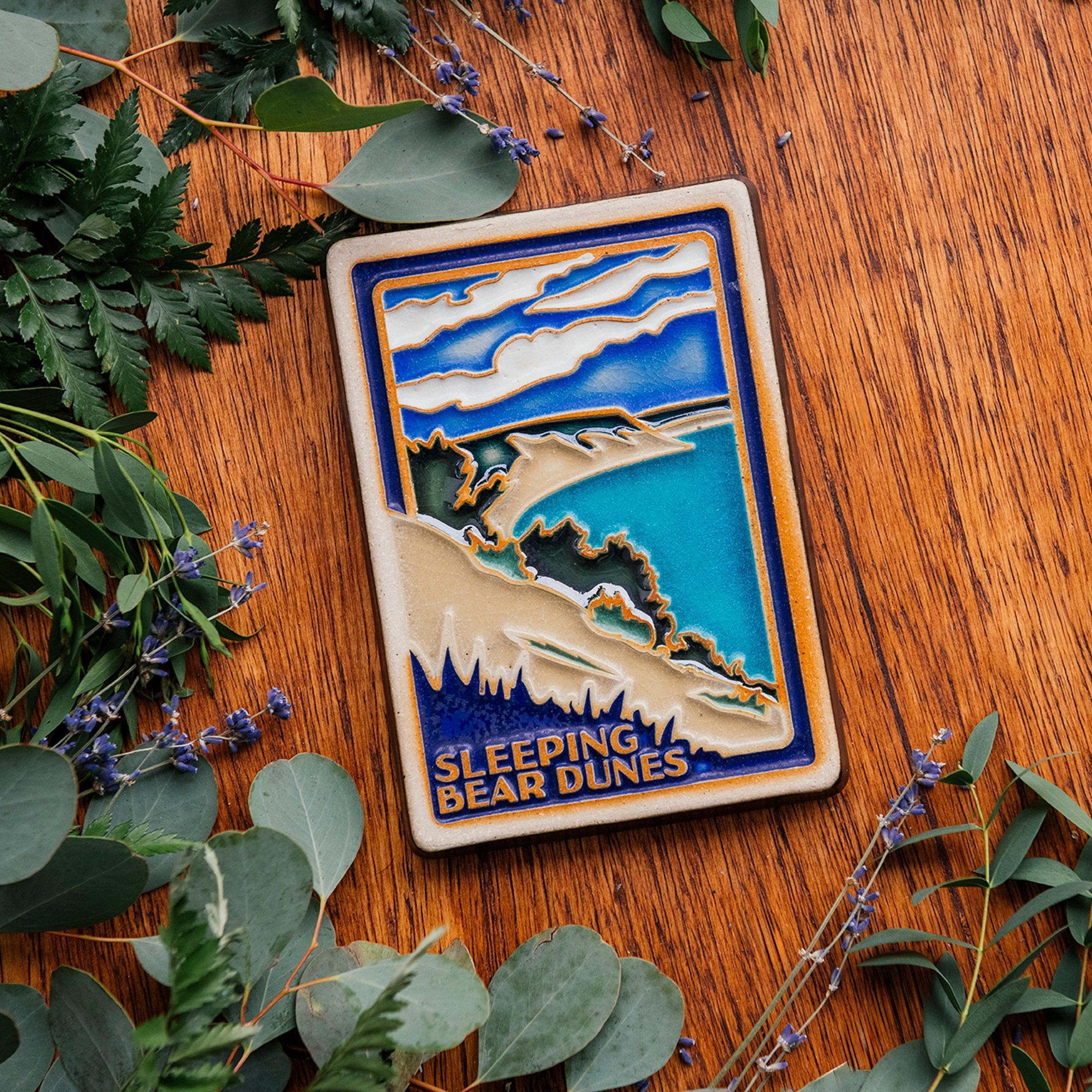 The Sleeping Bear Dunes Tile features the serene Lake Michigan and the sandy beaches and dunes covered on top by bushes and greenery. The words "Sleeping Bear Dunes" line the bottom of the design. The hand painted tile boasts blues and greens and pale yellow sand.