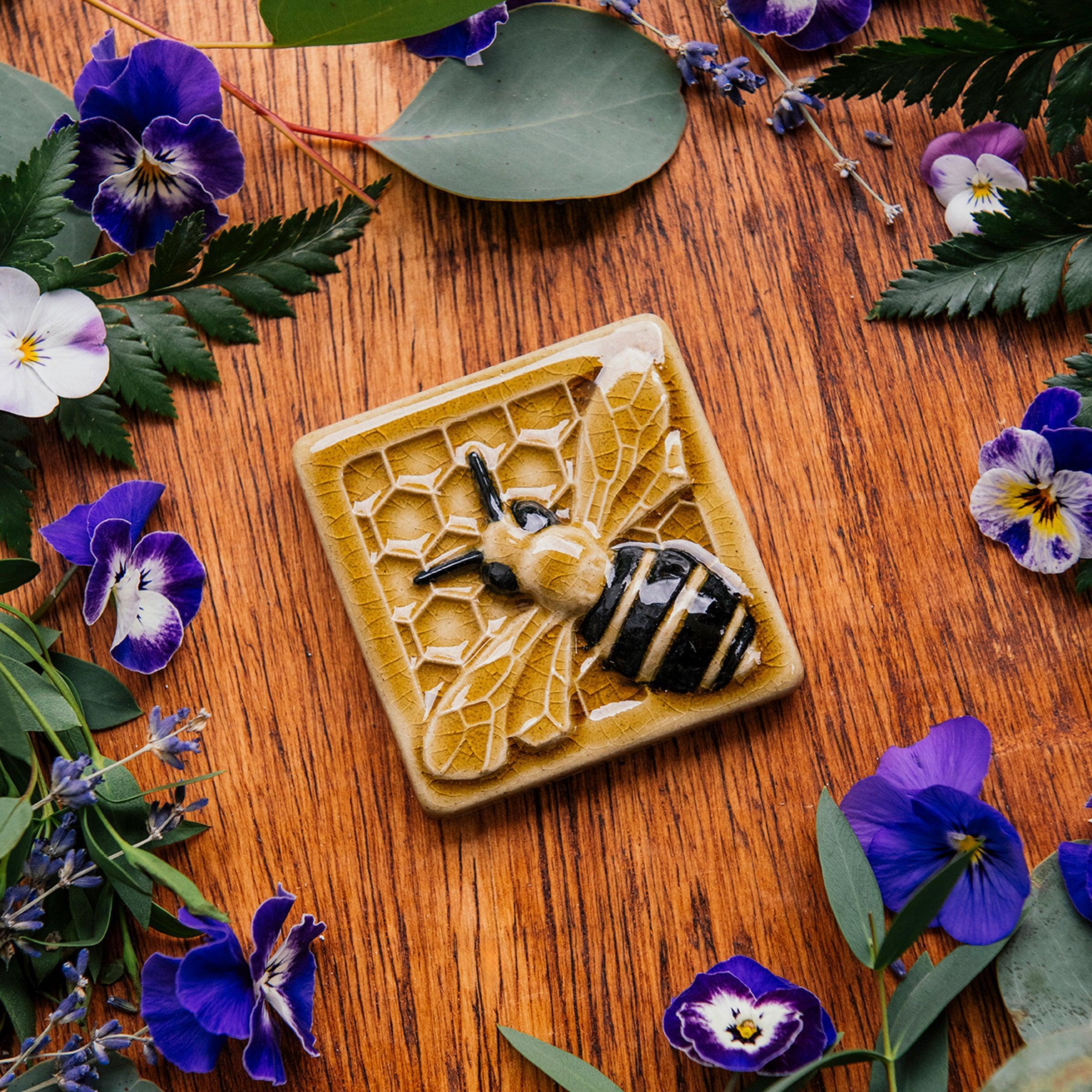 A glossy hand-painted Honeybee Tile in golden honey and black details surrounded by pansies, violets, and ferns. 