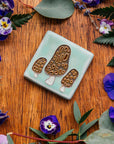 A tile depicting a line-drawn trio of brown Morel mushrooms on a light sea foam-glazed background. 