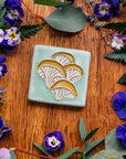 A hand-painted tile depicting a line drawing of Oyster mushrooms with golden yellow tops and white bases set on a light sea foam colored background glaze. 