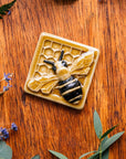 A glossy hand-painted Honeybee Tile in golden honey and black details surrounded lavender and summer greenery. 
