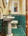 A wooden door opens to a custom tiled bathroom in varying greens and blues. The walls are an off-white color and the bathroom accents colors are mostly white and brass. 