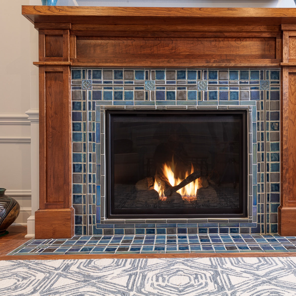A lively mix of blue, gray, and charcoal tiles are used in this fireplace and hearth set in a Craftsman style wooden mantel. There are two Pewabic Classic Vases on either side of the mantel in our true blue "Peacock" glaze. The fire is lit and there is a gray and white geometric rug in view on the floor.