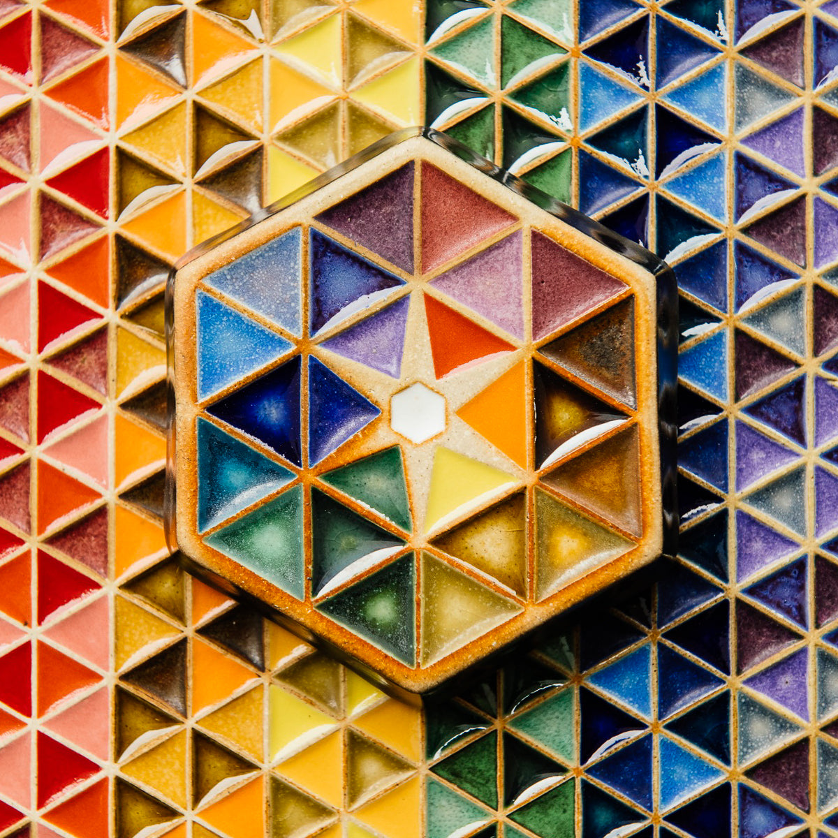 The Hex Paperweight is a thick ceramic tile shaped like a hexagon. The surface of the tile features an interlocking pattern of colorful triangles. In the center of the design is a small white hexagon with a starburst pattern exploding from it. This handpainted paperweight features 24 separate glazes, matte and glossy, in blues, greens, purples, yellows and oranges.