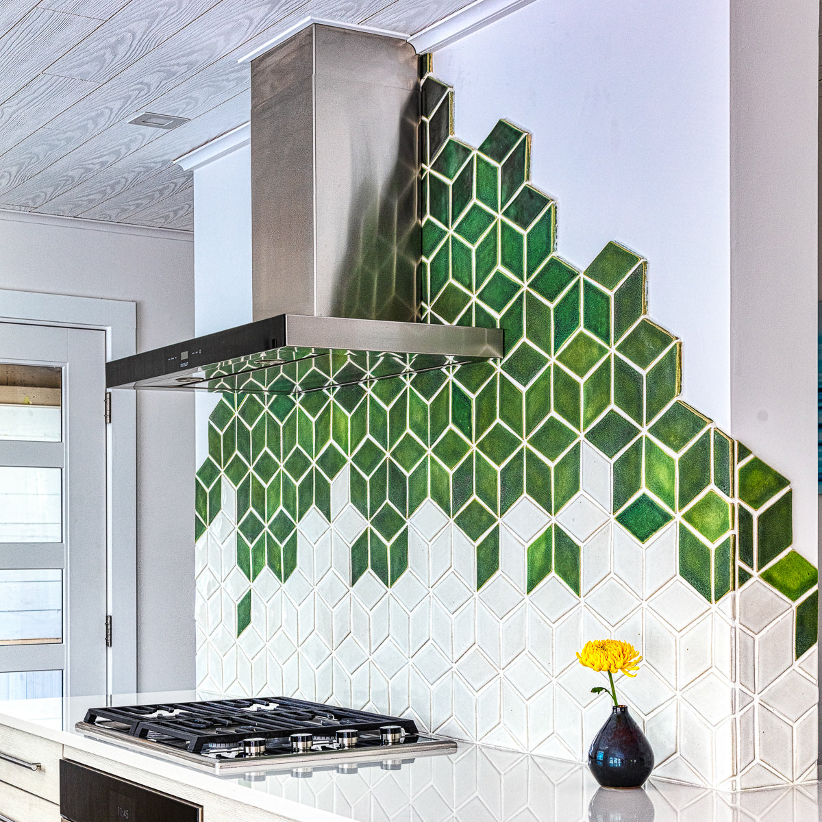 Modern kitchen with range hood against a custom tile backsplash comprised of 4" Rhombus tiles in a gradation of varying green and white glazes. There is a Pewabic Bud Vase with a single yellow flower resting on the glossy white countertop.