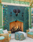 Large scale tile fireplace with high-gloss deep green and blue tiles. The fire is lit and there is a metal elephant head at the center of the design alongside two metal torches. There are light green pillars on each side of the fireplace. There is a wicker furniture set with sea-foam green cushions just within frame.  